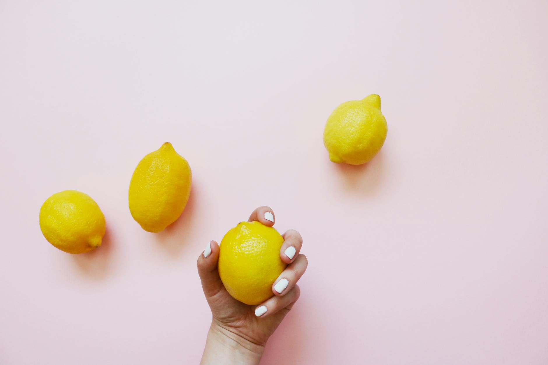 Una mano con unghie bianche tiene un limone su uno sfondo rosa tenue, circondata da altri limoni