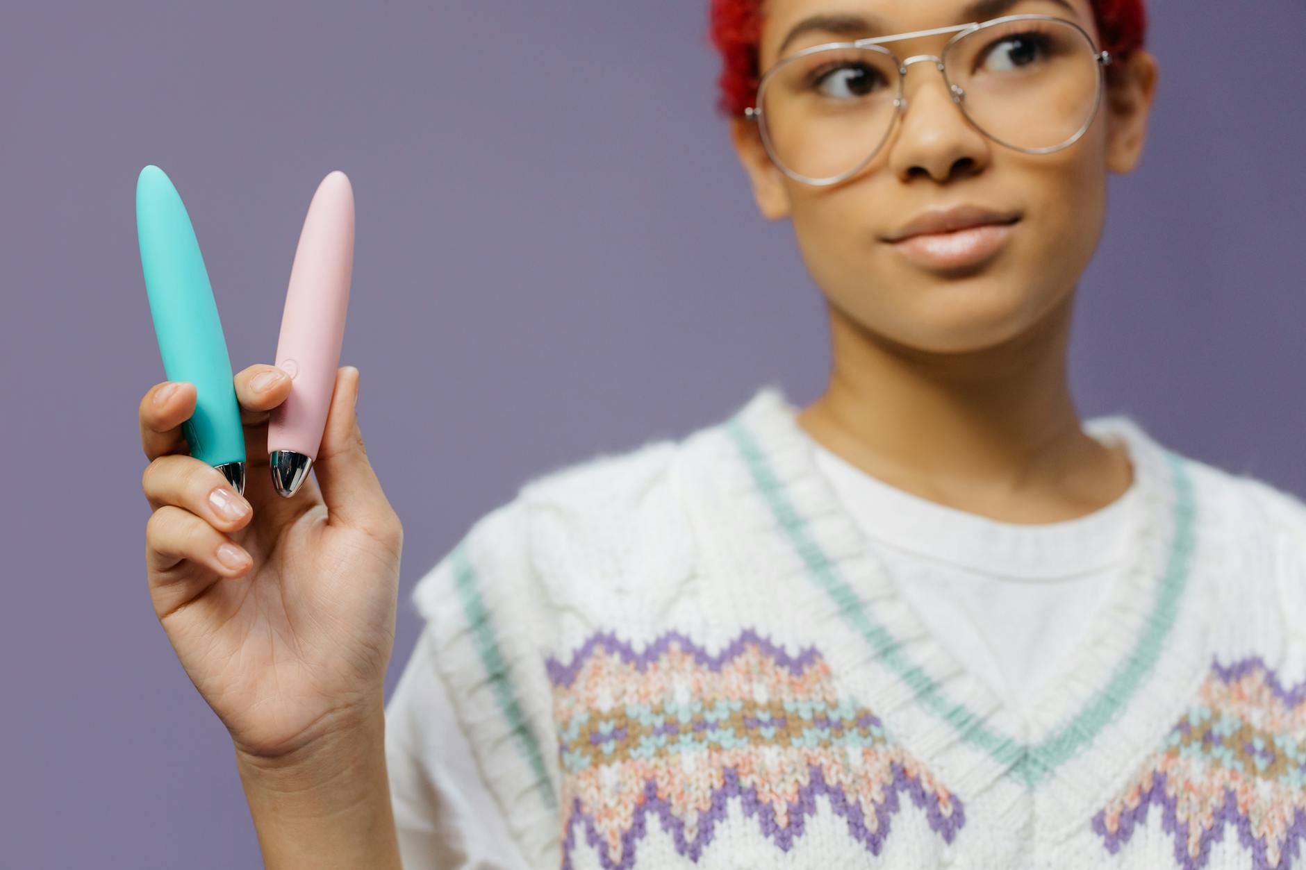 Una persona che indossa occhiali tiene due vibratori colorati contro uno sfondo viola
